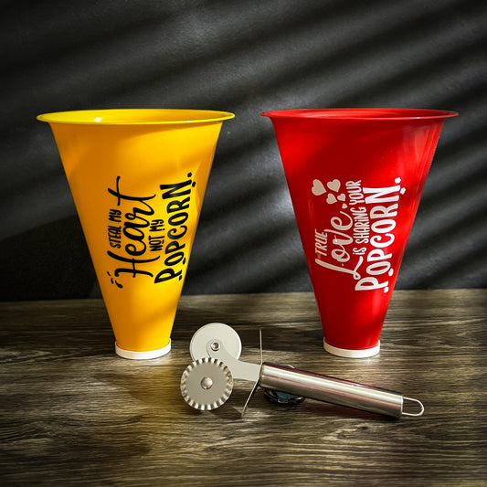 A set of popcorn containers labeled "His" (yellow) and "Hers" (red) with a sleek metal pizza cutter in front, displayed on a wooden surface.