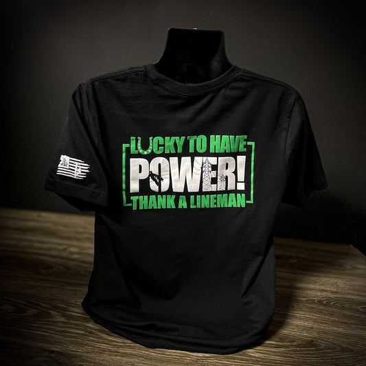 A black T-shirt featuring bold green and white text that reads "Lucky To Have Power! Thank A LineMan." The design emphasizes appreciation for LineMen, with the LineCrate logo and flag graphic on the sleeve. This shirt is crafted from durable cotton, offering both comfort and pride for those in the line trade.