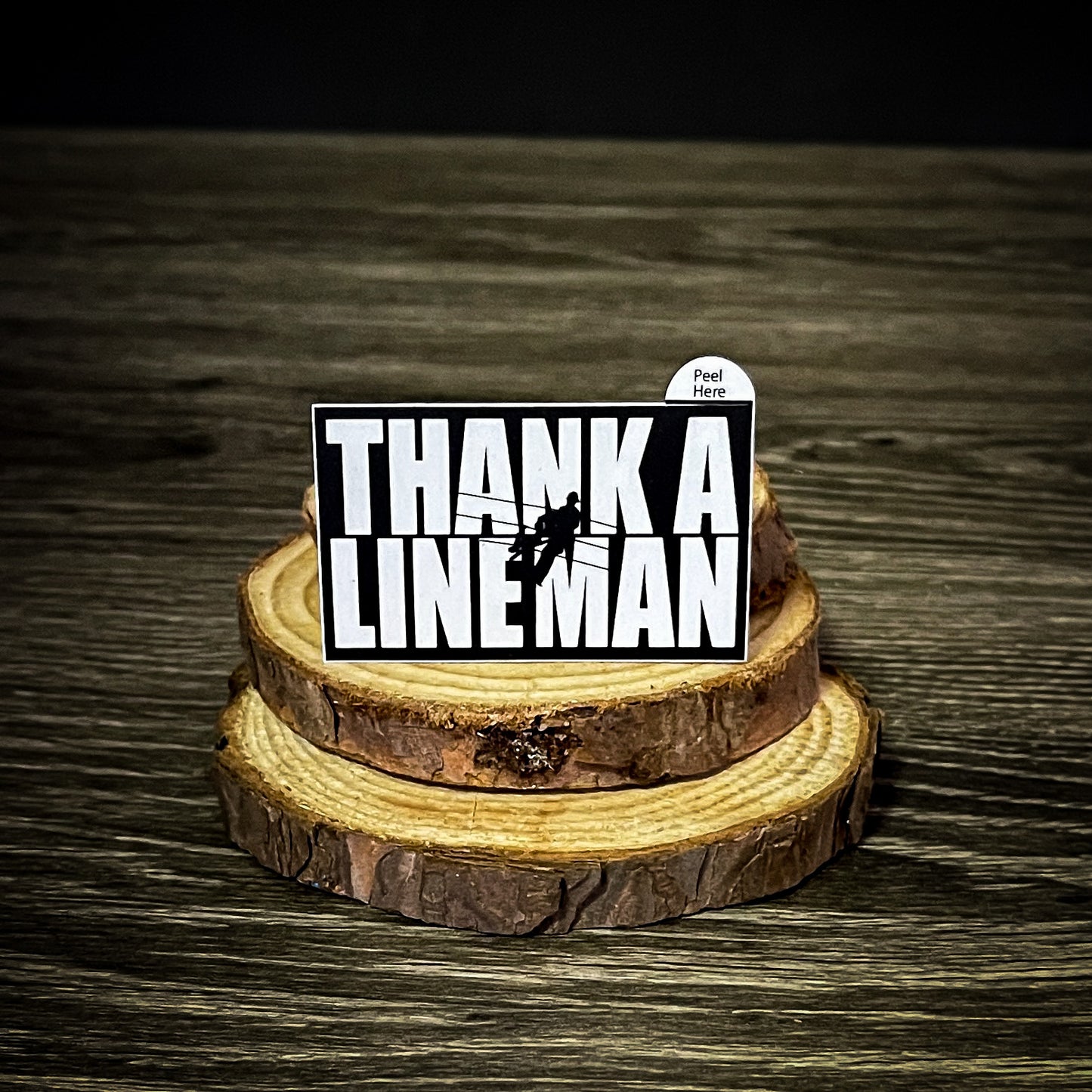 "Thank A LineMan" sticker featuring a black and white design with a lineman on a utility pole, displayed on a wooden surface. Size 2.5" x 1.5".