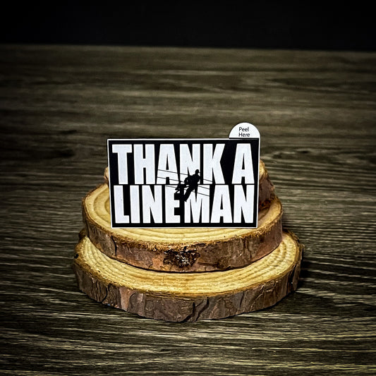 "Thank A LineMan" sticker featuring a black and white design with a lineman on a utility pole, displayed on a wooden surface. Size 2.5" x 1.5".
