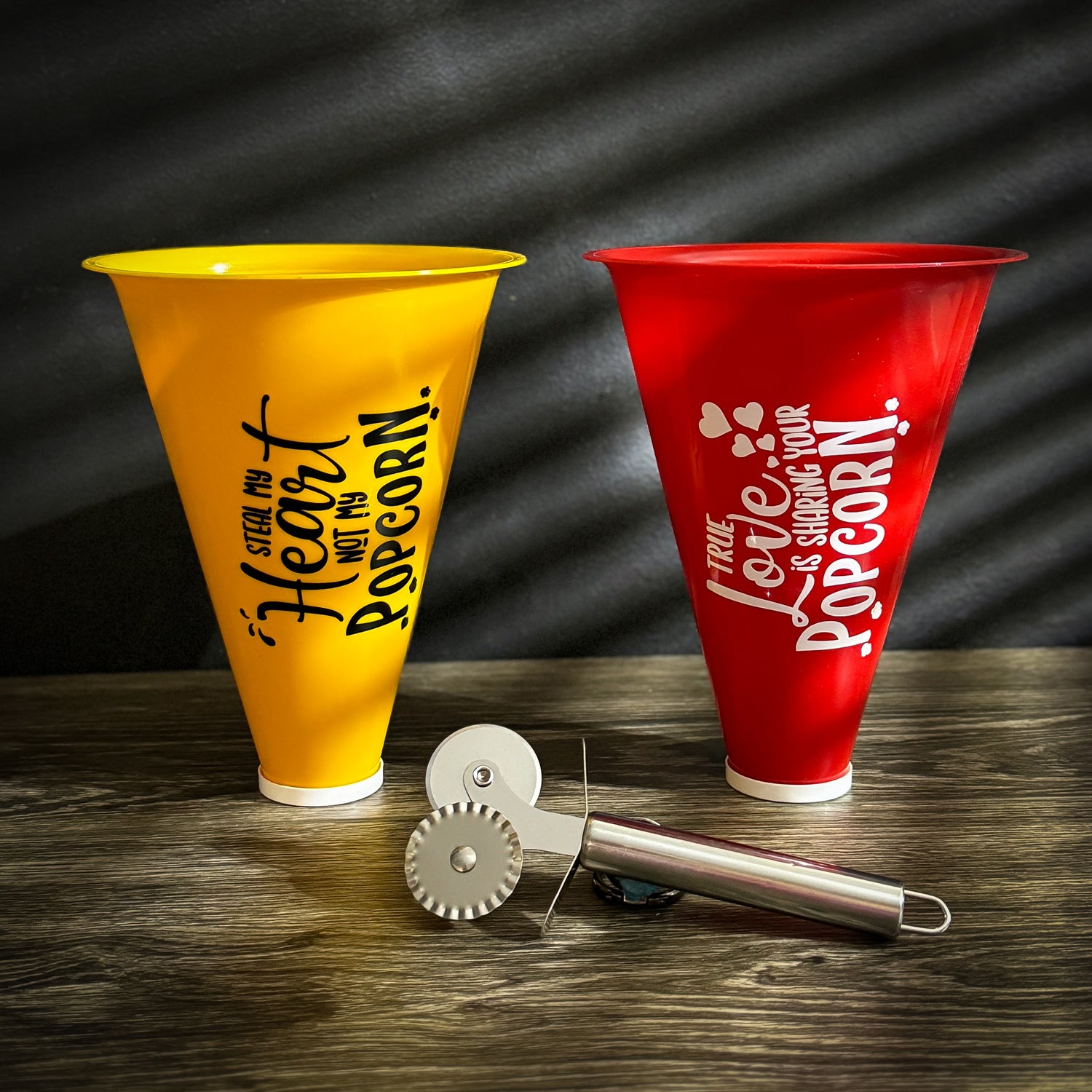  A set of popcorn containers labeled "His" (yellow) and "Hers" (red) with a sleek metal pizza cutter in front, displayed on a wooden surface.