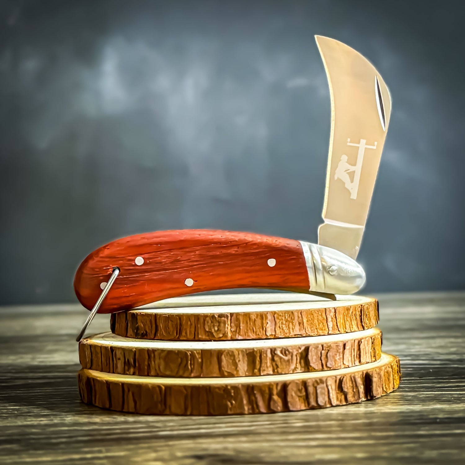 A hawk bill wooden handle and a stainless-steel engraved with a lineman silhouette, displayed on a wood-stacked stand.