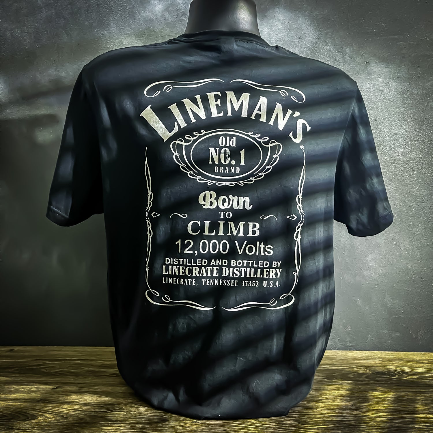 Black t-shirt with a whiskey label-inspired design that reads "LineMan's Born to Climb 12,000 Volts" in bold white letters. Features LineCrate logo on the sleeve.