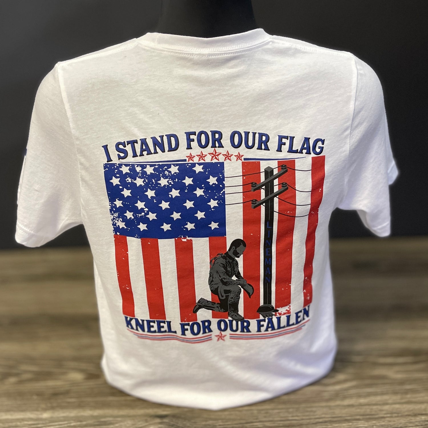 White t-shirt featuring a large American flag graphic with a kneeling blue collar man and the words "I Stand for Our Flag, Kneel for Our Fallen.