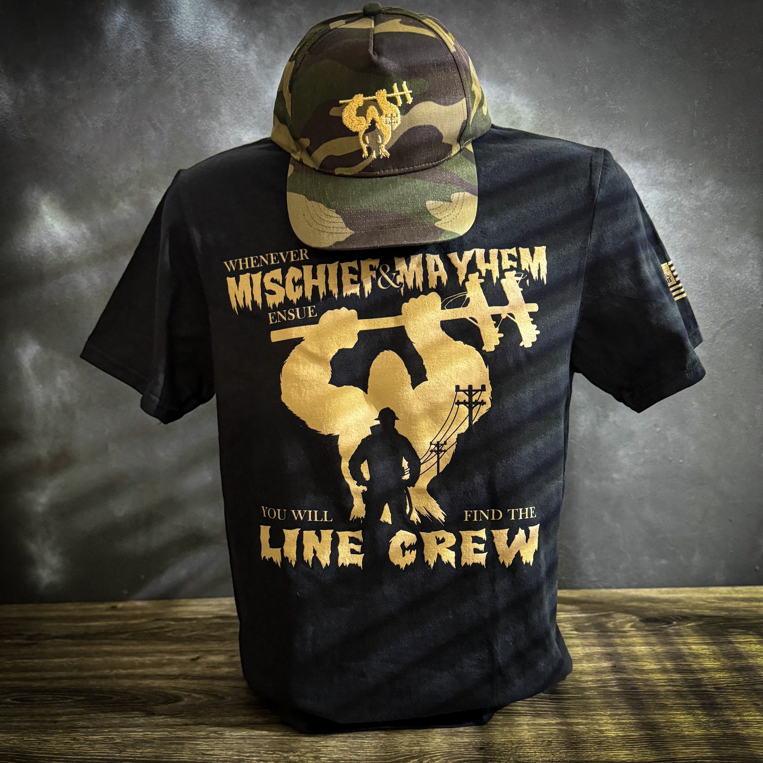 Black t-shirt with yellow lettering that says, "Whenever Mischief & Mayhem Ensue, You Will Find Line Crew," featuring a silhouette of a LineMan and utility poles in the background. The sleeve displays the LineCrate flag logo with a LineMan on a pole.