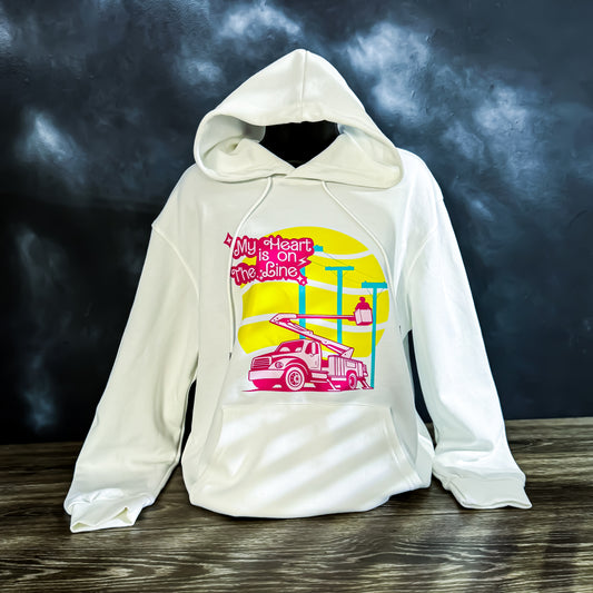 White hoodie with a colorful graphic of a lineman working on power lines and the phrase "My Heart is on the Line" printed in pink, yellow, and teal.