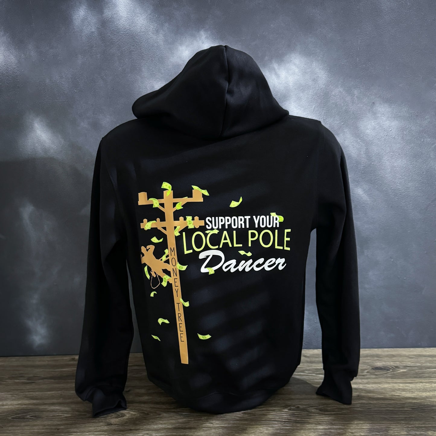 Black hoodie featuring a graphic of a lineman climbing a utility pole, labeled as a "money tree" with dollar bills flying around. The phrase "Support Your Local Pole Dancer" is printed in bold white and green lettering, making it a humorous tribute to hardworking LineMen.