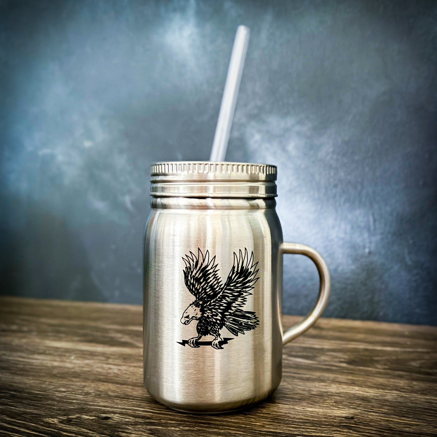 Stainless steel mason jar with black eagle design and attached handle, featuring a lid and clear straw